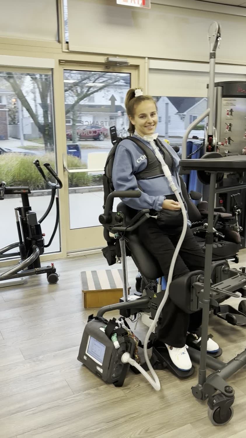 Rebecca smiling, supported upright in a powered standing frame during rehab.