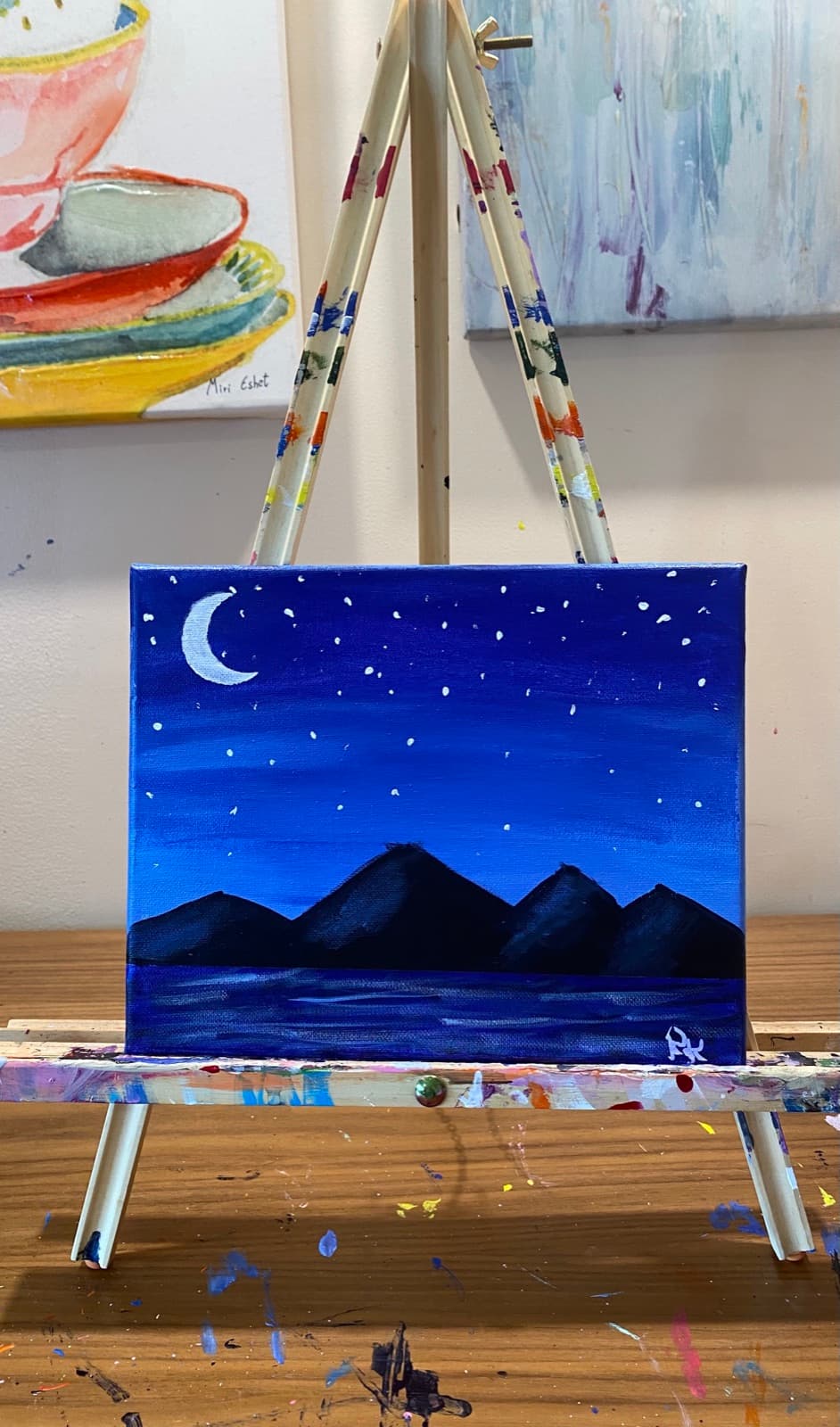 Mouth painting: navy night sky with a crescent moon over dark mountains and water.