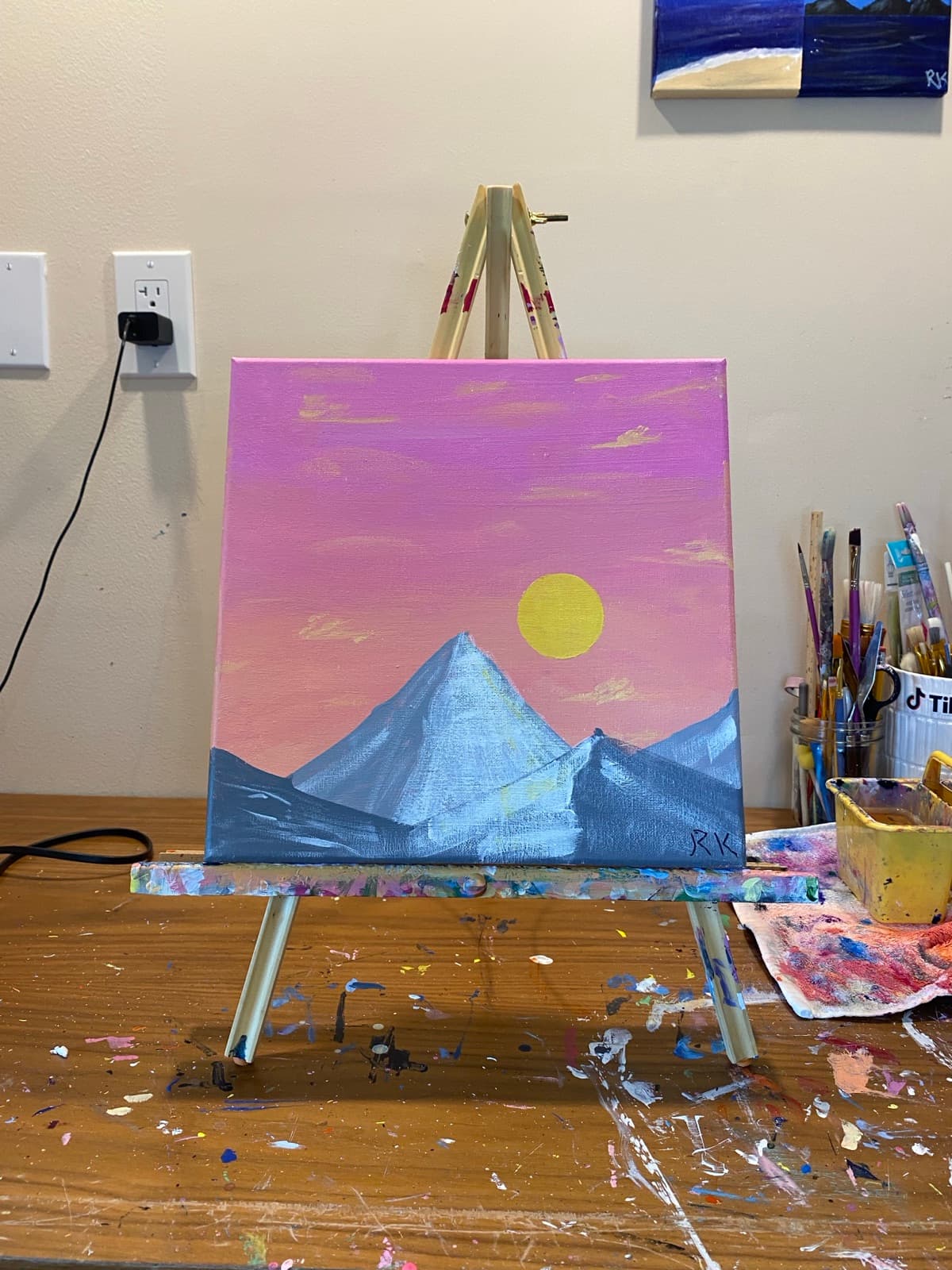 Mouth painting: a pink and yellow sky with a sun above purple and blue mountains.