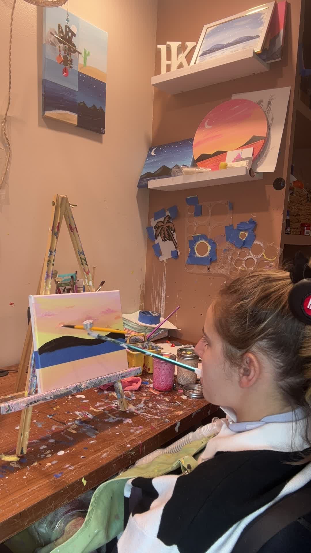 Rebecca painting at her easel, holding a brush in her mouth, surrounded by her finished landscape paintings.